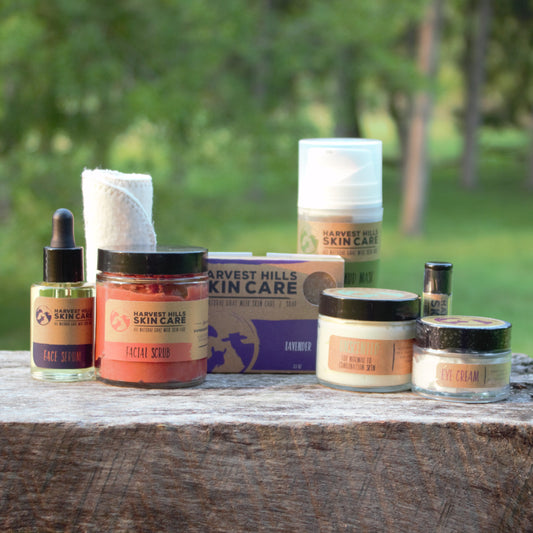 A collection of Harvest Hills Skin Care products arranged on a rustic wooden surface outdoors with a blurred green forest background. The products include a face serum, facial scrub, Mud Mask, face cream, lip balm and an eye cream, with a organic cotton face cloth and bar of goat milk soap behind them.