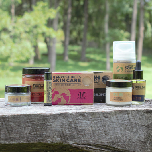A set of Harvest Hills Skin Care face care products for combination skin arranged on a rustic wooden ledge with a blurred forest background. The products include two bars of goat milk soap, a lip balm tube, face serum, clarifying mud mask and eye cream 