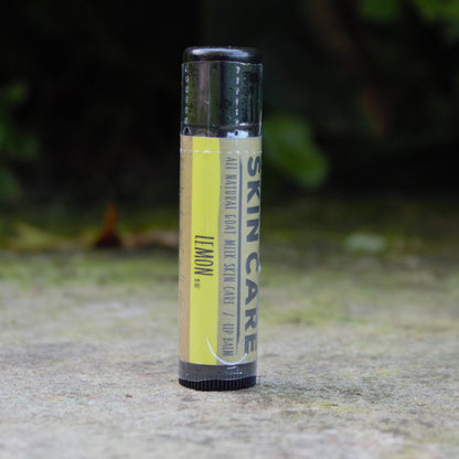 A single tube of Harvest Hills SKIN CARE lip balm, Lemon, standing upright on a concrete surface with a blurred green garden background.
