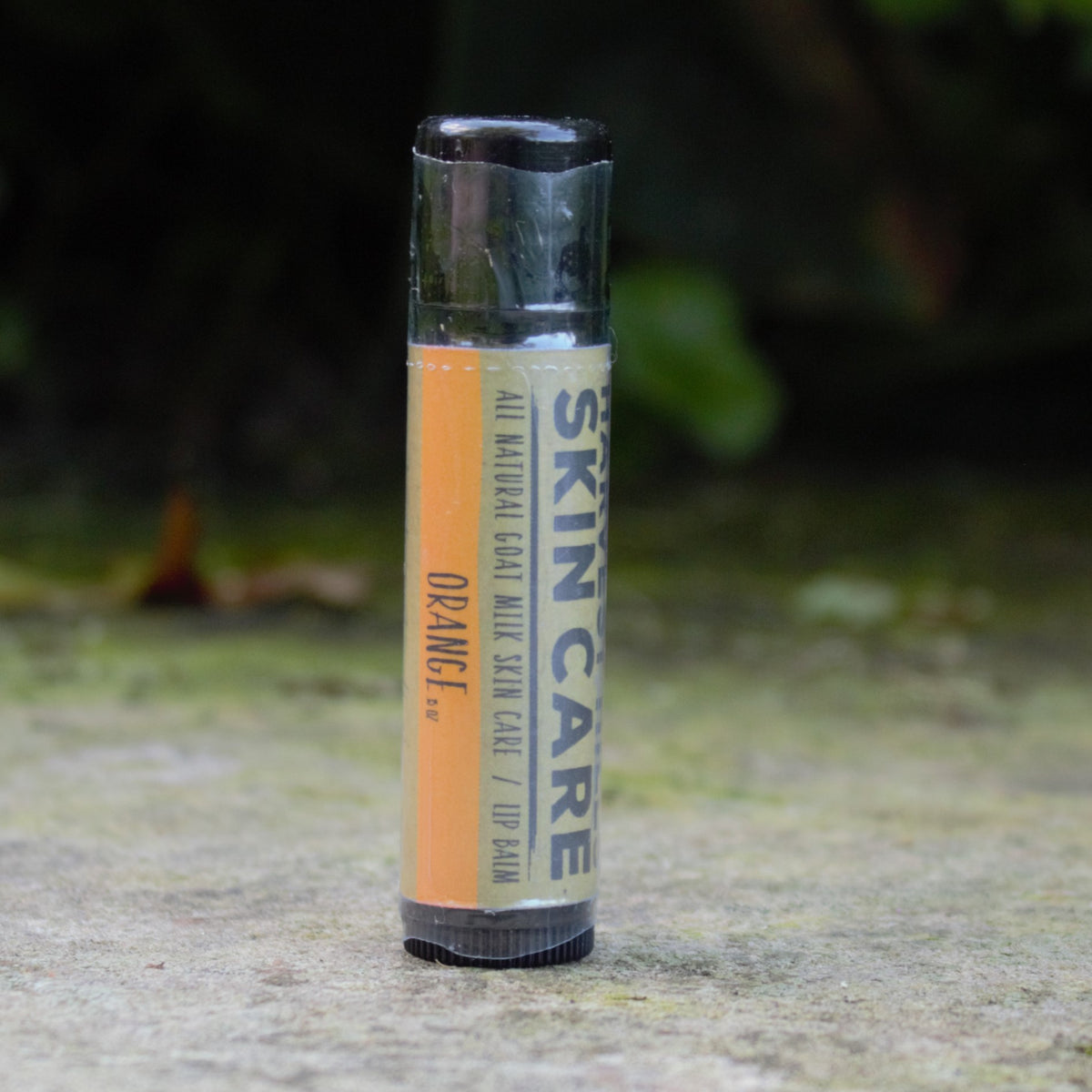 A single tube of Harvest Hills Skincare Orange lip balm standing upright on a stone surface with a blurred natural green background.